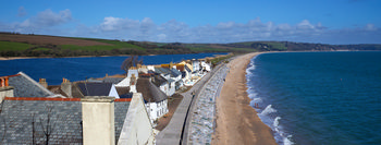 Slapton sands beach from above The image is a landscape photograph of Slapton Sands beach on the coast of Devon, England, in the United Kingdom. The scene shows the long, sweeping stretch of Slapton Sands beach from an elevated viewpoint, likely taken during the late morning in early spring. To the left, traditional houses with slate rooftops line the edge of the village, while a narrow road runs parallel to the shoreline, separating the dwellings from the sand. The beach extends into the distance, bordered by the vivid blue sea on one side and the freshwater lake of Slapton Ley on the other, with rolling green hills visible further inland. The clear sky suggests a bright spring day, adding clarity to the coastal features and the rural landscape of this region in Devon, England.